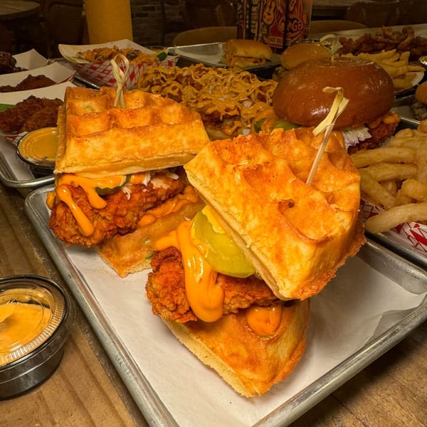 Two Flamin Feathers fried chicken and waffle sandwiches with pickles and sauce, surrounded by other dishes.