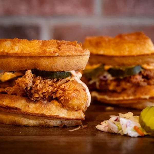 Two Flamin Feathers fried chicken and waffle sandwiches with pickles and coleslaw on a wooden surface.