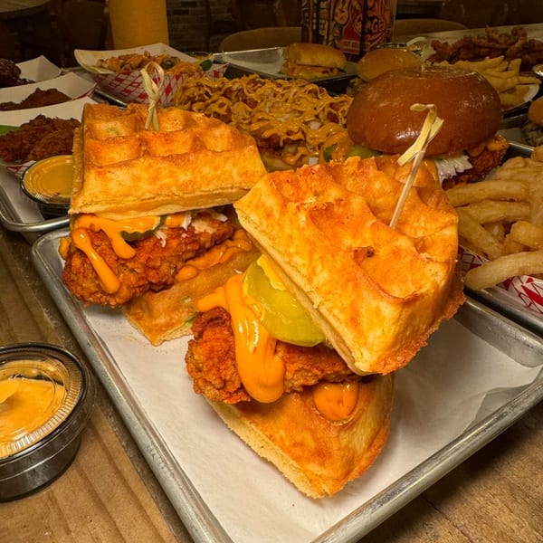 Two Flamin Feathers fried chicken and waffle sandwiches with sauce and pickles, surrounded by other fried chicken dishes.