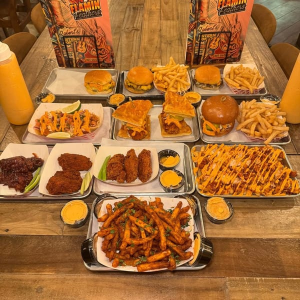 A wooden table laden with various Flamin Feathers fried chicken dishes, fries, burgers, and sauces.